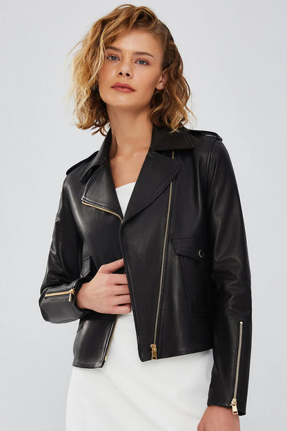 Woman styling the black asymmetrical biker leather jacket open over a white dress, showing the cropped hem and drape of the lambskin leather.